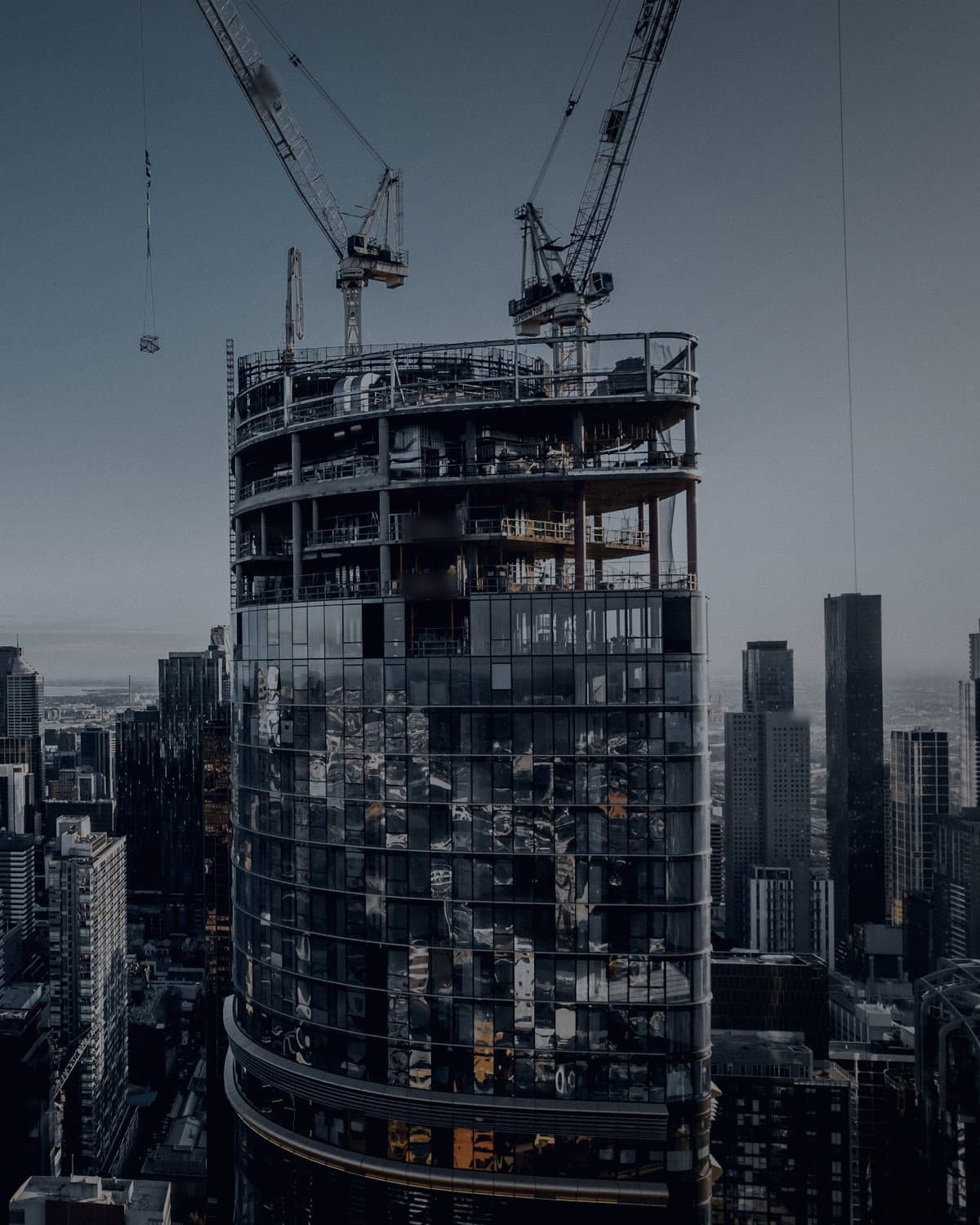 Two tower cranes above a curved tower under construction, the city behind it in early light.